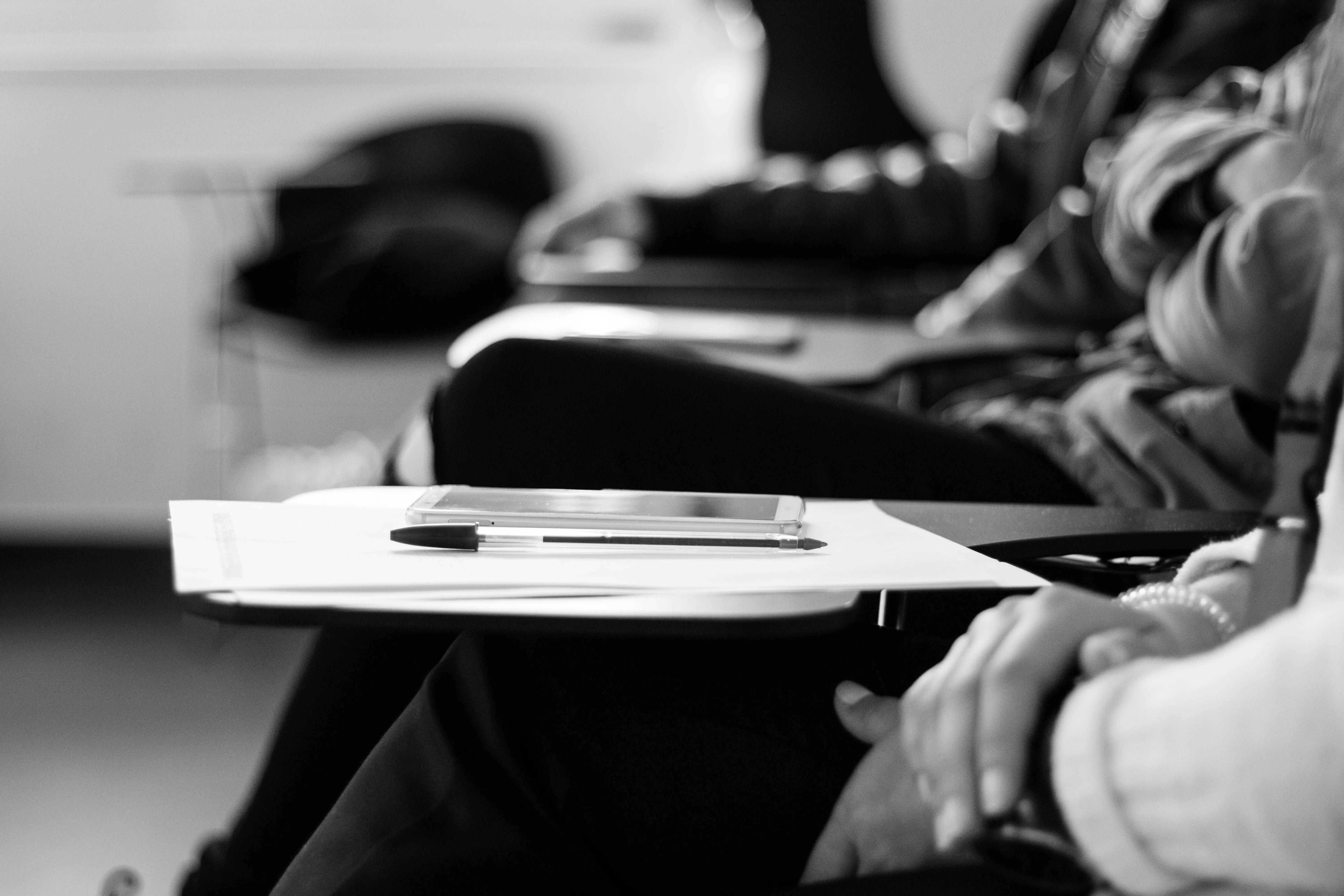 Grayscale photo of students sitting on chairs with papers and pens